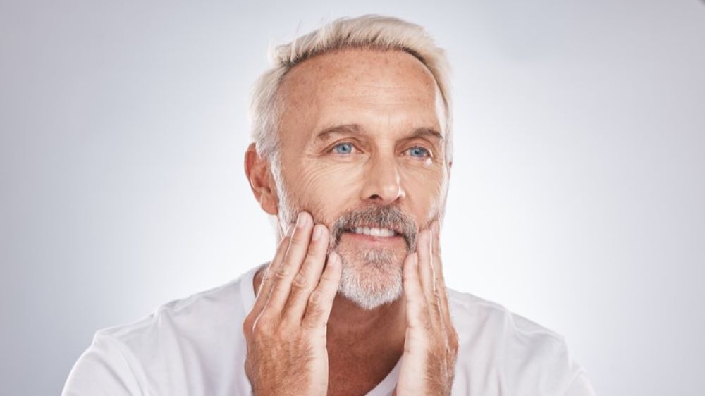 A mature man with white hair and a beard gently touches his face, displaying a thoughtful expression.
