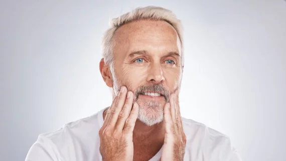A mature man with white hair and a beard gently touches his face, displaying a thoughtful expression.